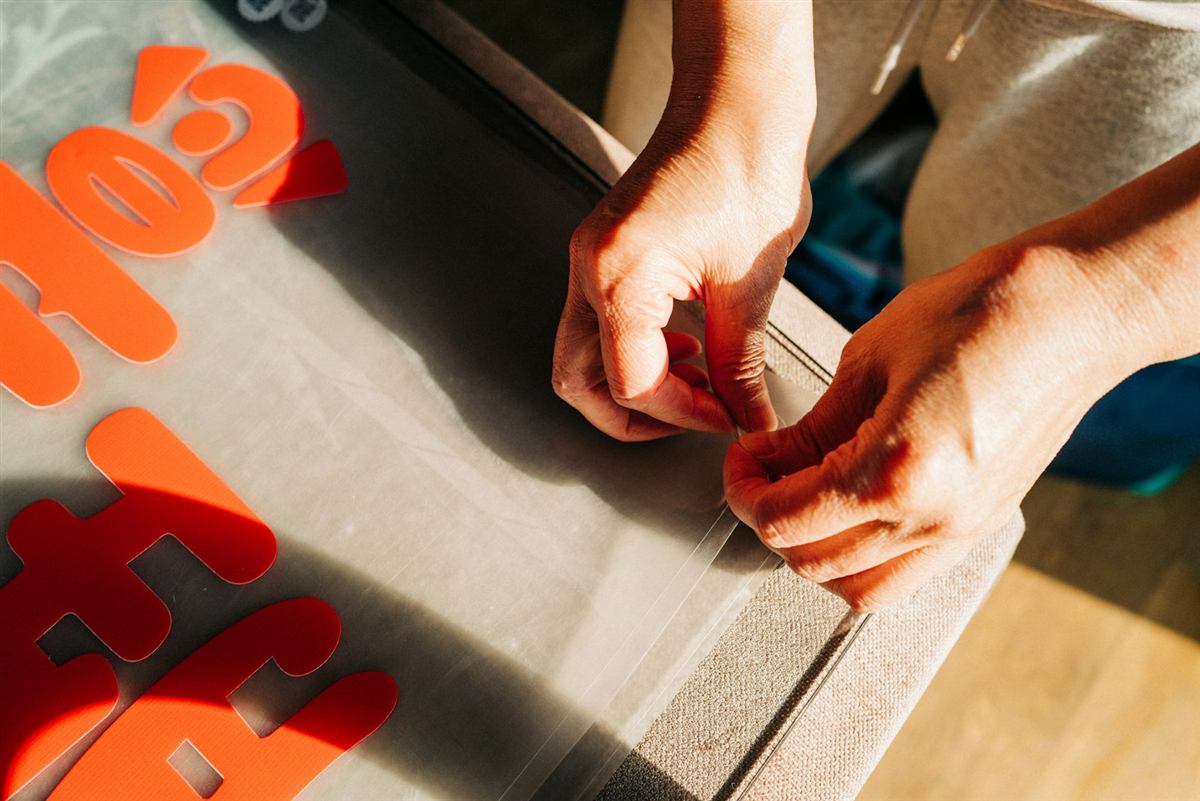 Hände lösen eine Folie vom Trägerpapier, daneben orange Folienbuchstaben